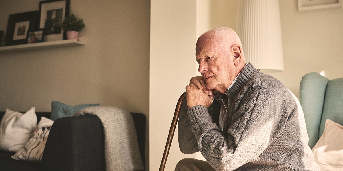 An older man in his home leaning on a walking stick 